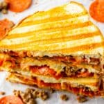 Stack of half pizza panini sandwiches with pepperoni and sausage scattered on table