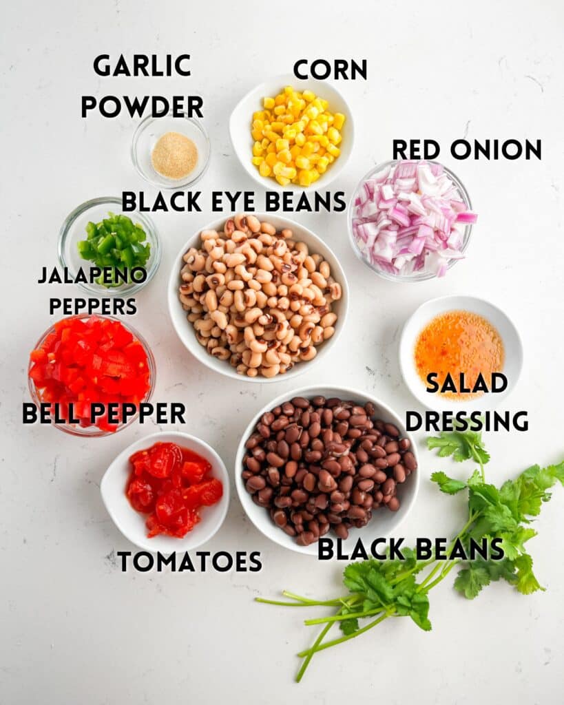 Top-down view of labeled salad ingredients for Cowboy Caviar, including black beans, black eye beans, corn, red onion, bell pepper, tomatoes, jalapeño peppers, garlic powder, salad dressing, and cilantro on a white surface.