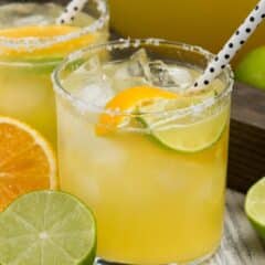 yellow drink in a small clear glass with sliced limes and oranges and a spotted straw.