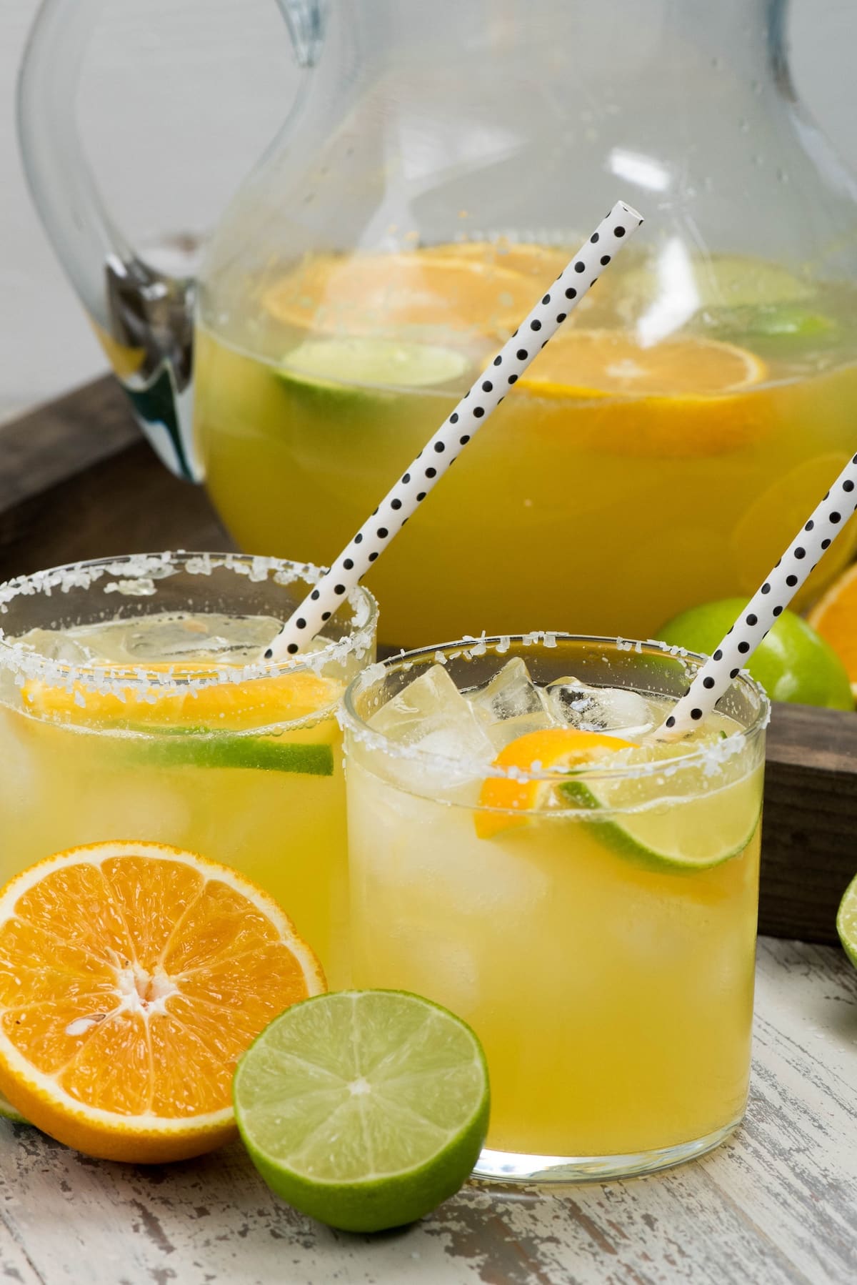 yellow drink in a small clear glass with sliced limes and oranges and a spotted straw.