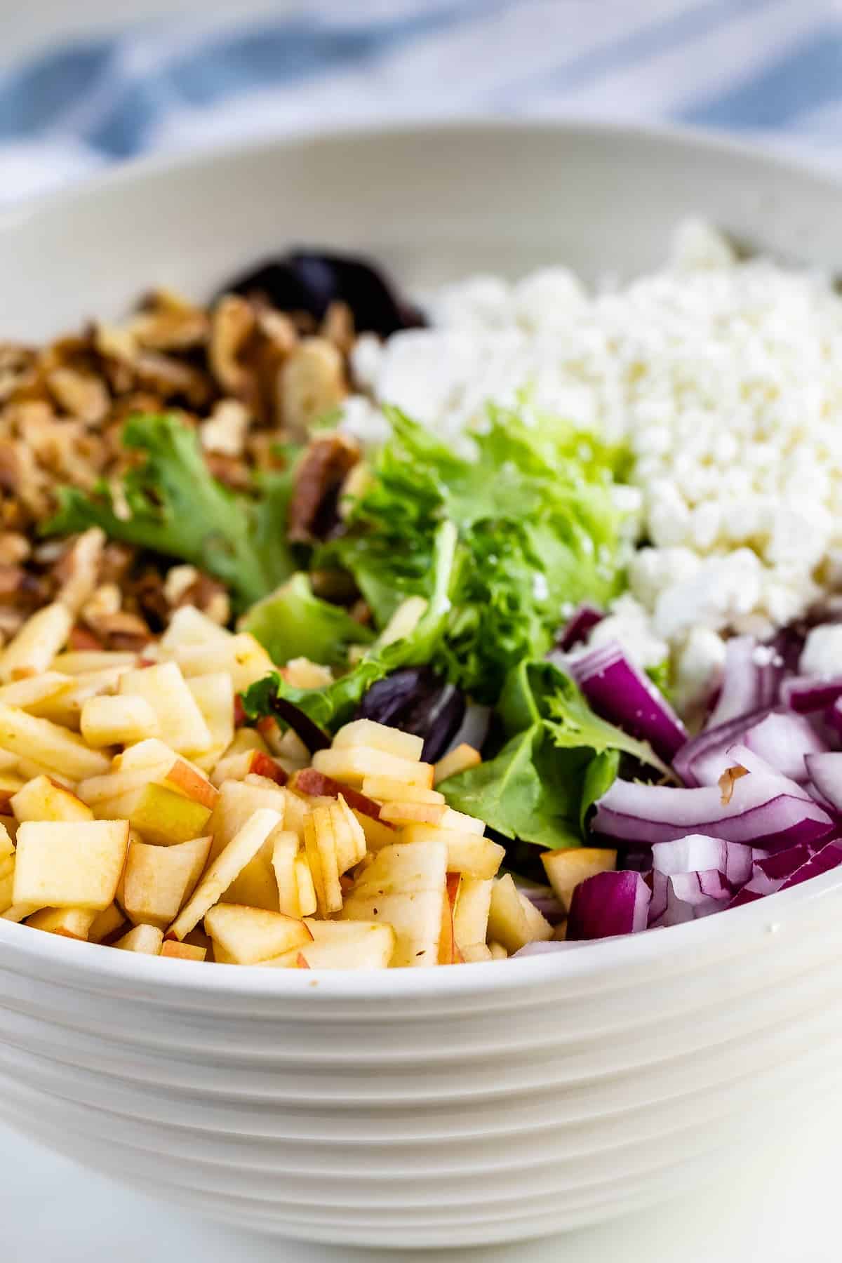 apples and walnuts and greens in a white bowl next to a wooden spoon.