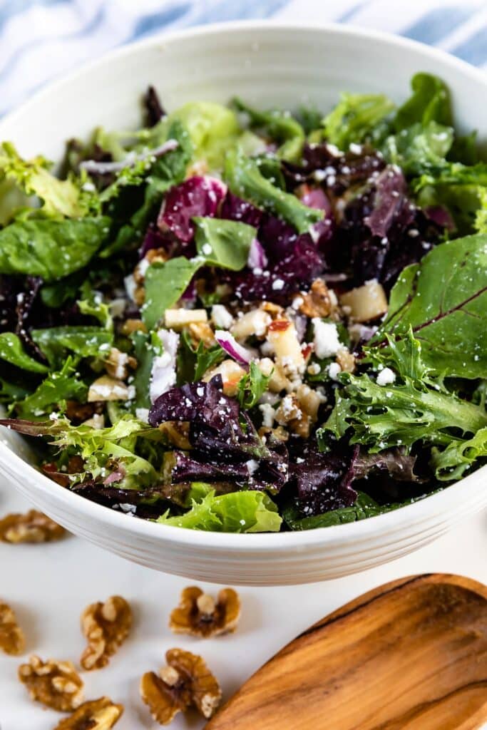 apples and walnuts and greens in a white bowl next to a wooden spoon.