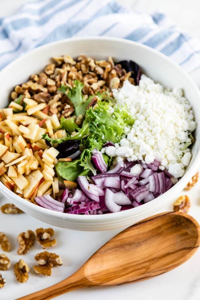 apples and walnuts and greens in a white bowl next to a wooden spoon.