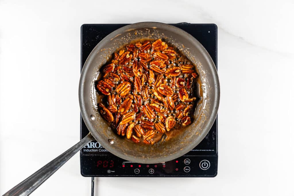 Top view of a frying pan on an induction cooktop, filled with candied nuts. The caramelized pecans are coated in a glossy sauce and are evenly distributed across the pan.