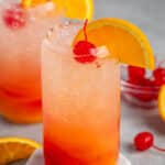 red and orange drink in a clear glass with an orange slice on the rim and a cherry on top.