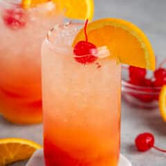 red and orange drink in a clear glass with an orange slice on the rim and a cherry on top.