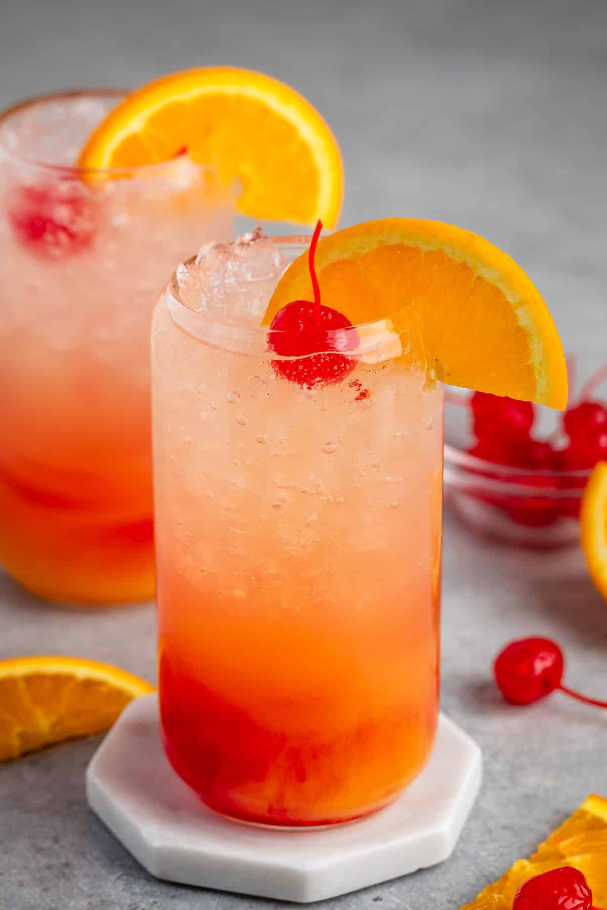 red and orange drink in a clear glass with an orange slice on the rim and a cherry on top.