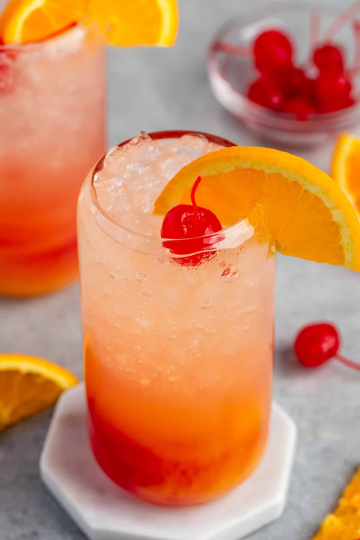 red and orange drink in a clear glass with an orange slice on the rim and a cherry on top.