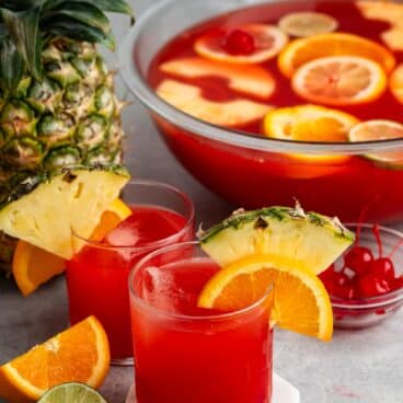 short clear glass with red drink inside and an orange and pineapple slice on the rim.