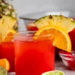 short clear glass with red drink inside and an orange and pineapple slice on the rim.