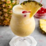 yellow Pina colada in a clear glass with a pineapple and cherry on the rim.