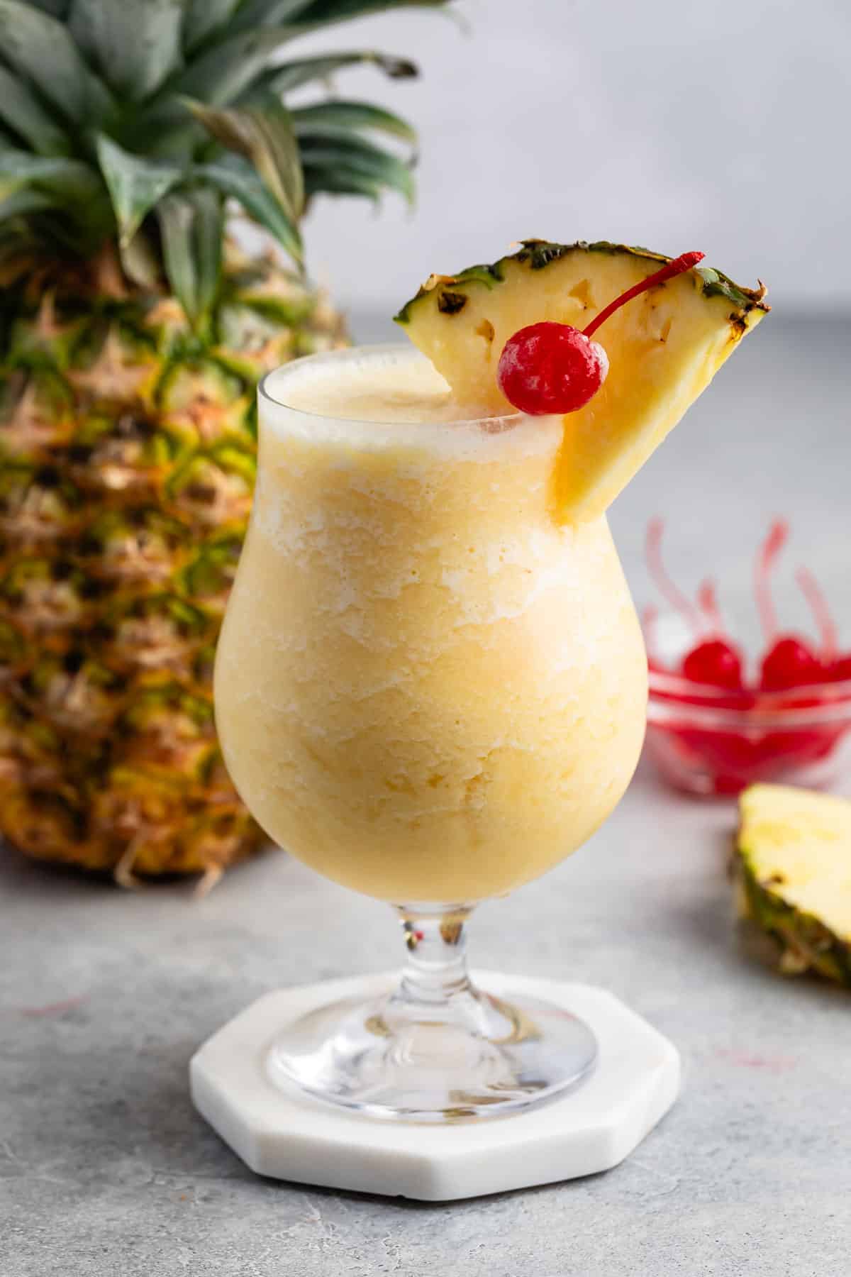 yellow Pina colada in a clear glass with a pineapple and cherry on the rim.