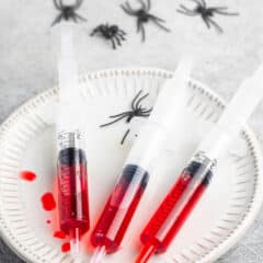 red grenedine in a plastic syringe laid out on a white plate next to fake spiders.