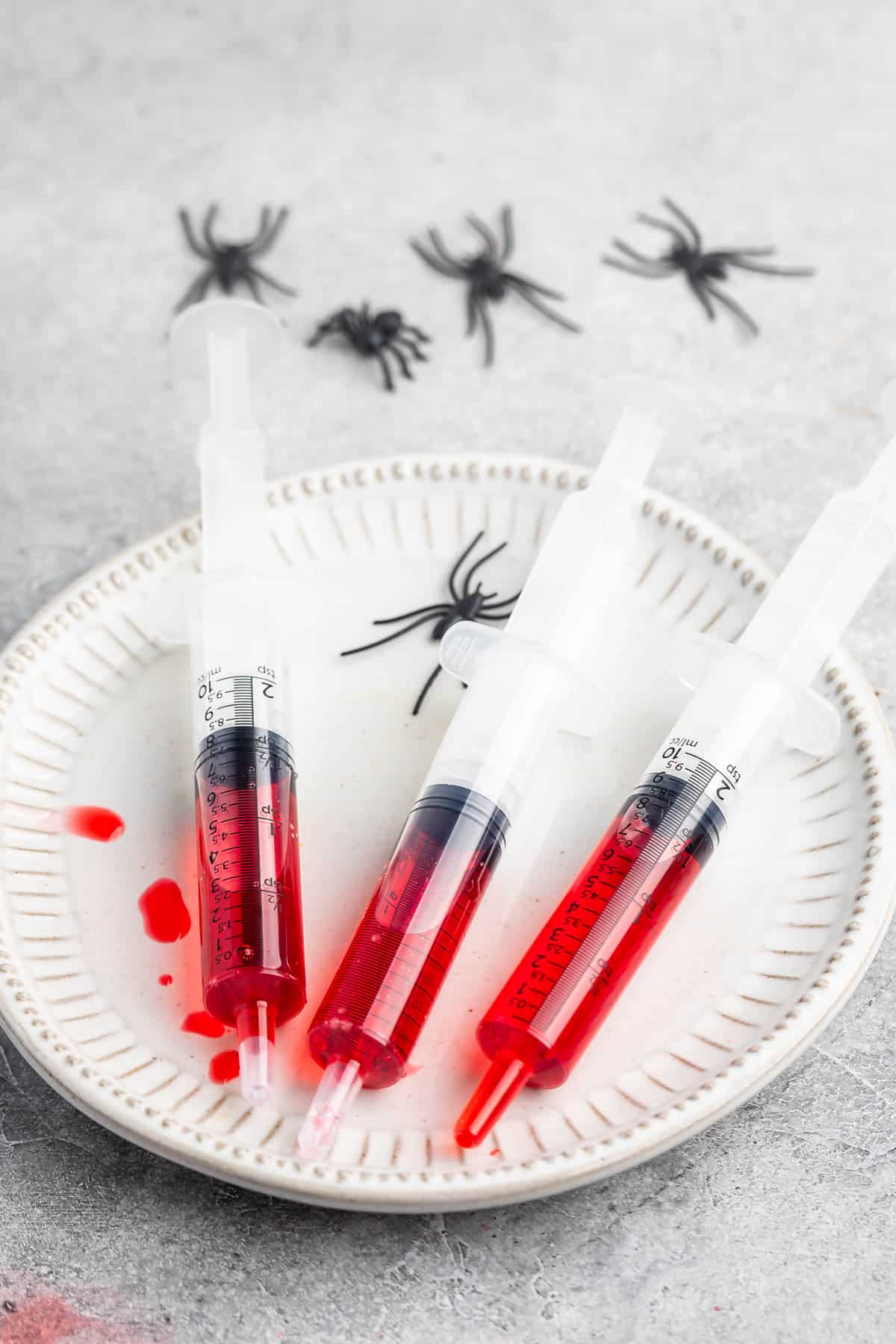 red grenedine in a plastic syringe laid out on a white plate next to fake spiders.