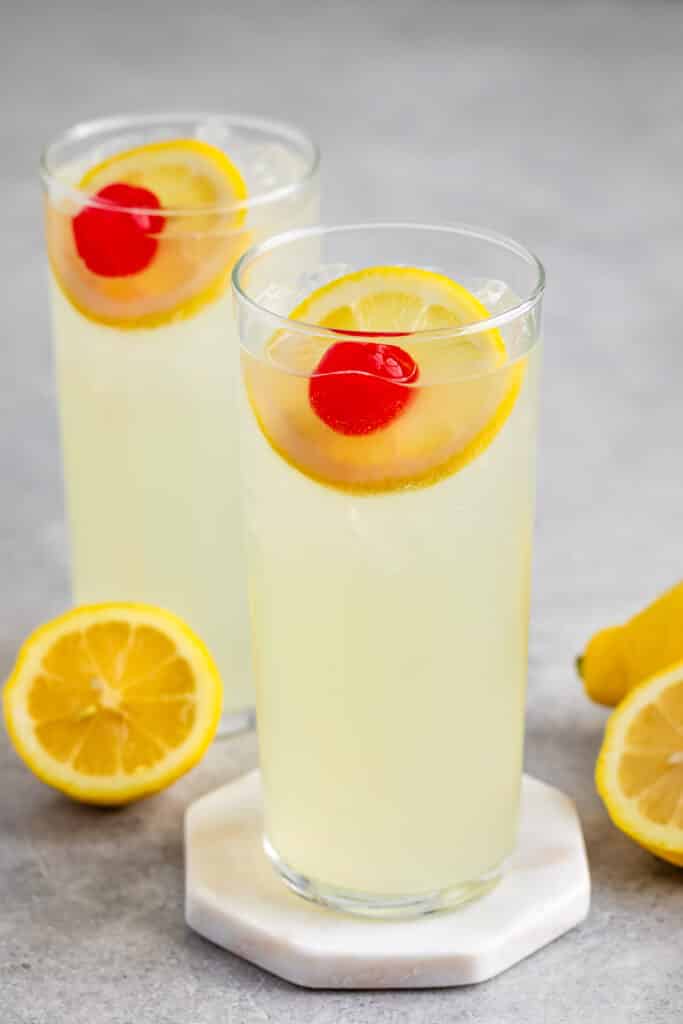 yellow drink mixed with a lemon slice and cherry in a tall clear glass.