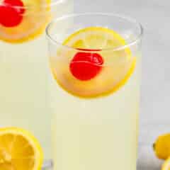 yellow drink mixed with a lemon slice and cherry in a tall clear glass.