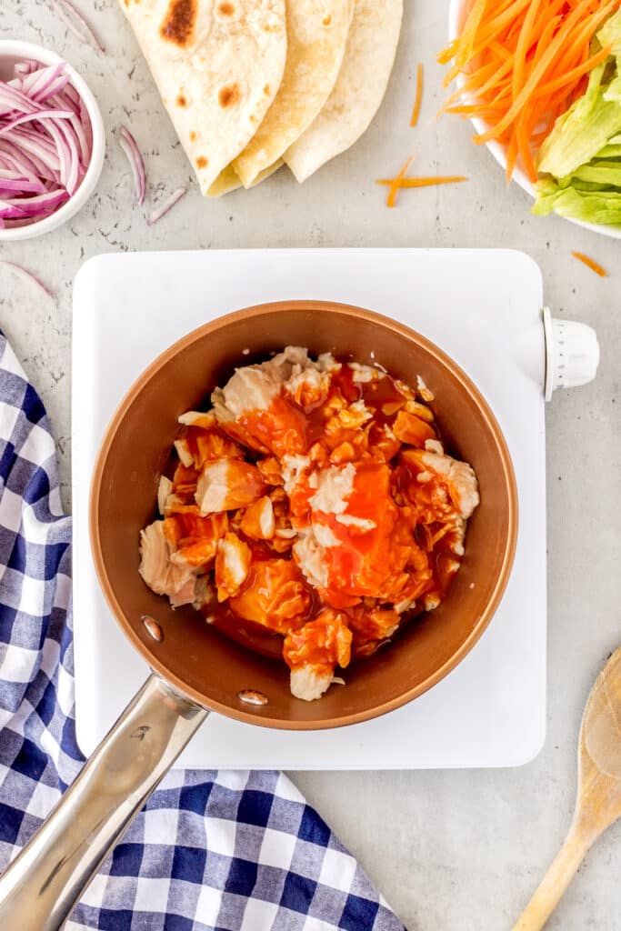 A pan on a white stovetop holds chopped chicken and red sauce. Surrounding the pan are plates with tortillas, sliced carrots, shredded lettuce, and a bowl of sliced red onions. A wooden spoon and blue checkered cloth are nearby.