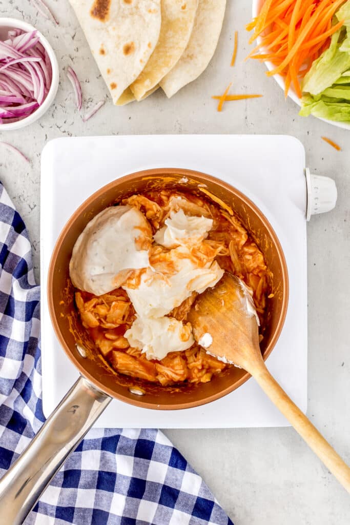 A skillet on a stovetop contains shredded chicken mixed with a creamy sauce. A wooden spoon is in the skillet. Surrounding the skillet are tortilla wraps, julienned carrots, lettuce, sliced red onions, and a checkered cloth.