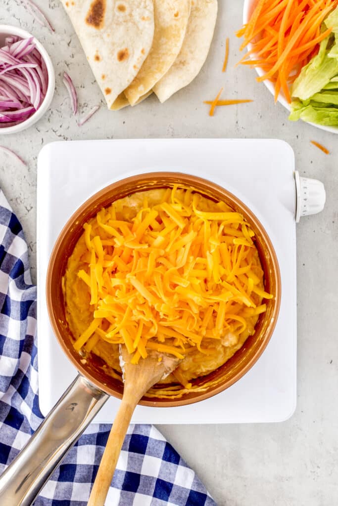 A pot of melted cheese on a stovetop, being stirred with a wooden spoon. Shredded carrots, tortilla wraps, lettuce, and red onion are nearby on a light gray surface, alongside a blue checkered cloth.