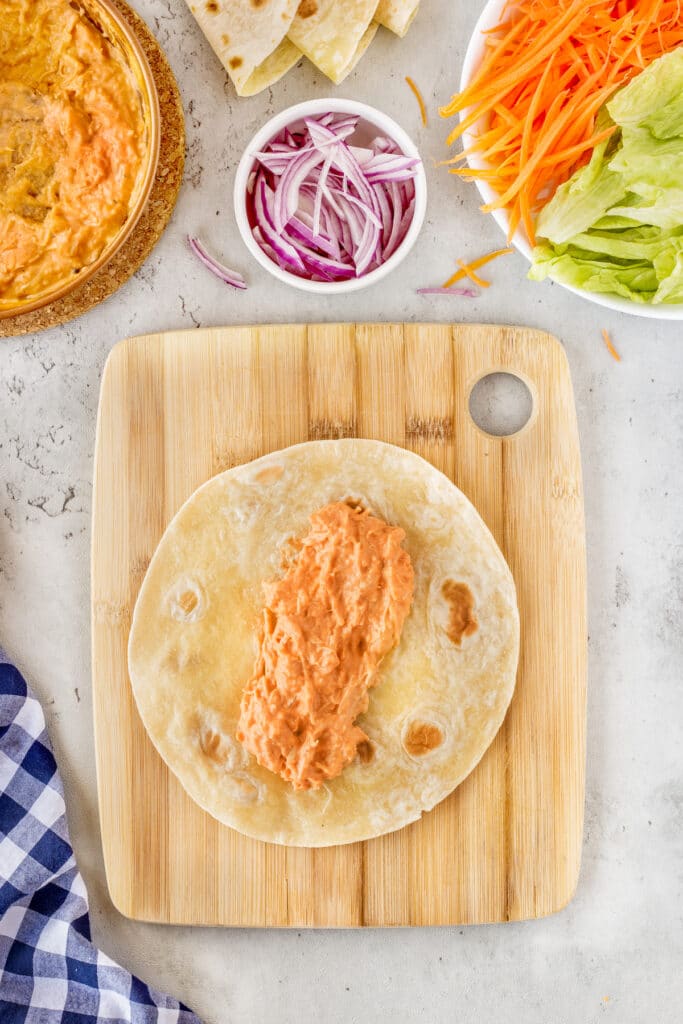 A wooden board holds a tortilla topped with a creamy orange spread. Surrounding it are sliced red onions, shredded carrots, lettuce, and extra flatbreads. A blue and white checkered cloth is partially visible to the side.