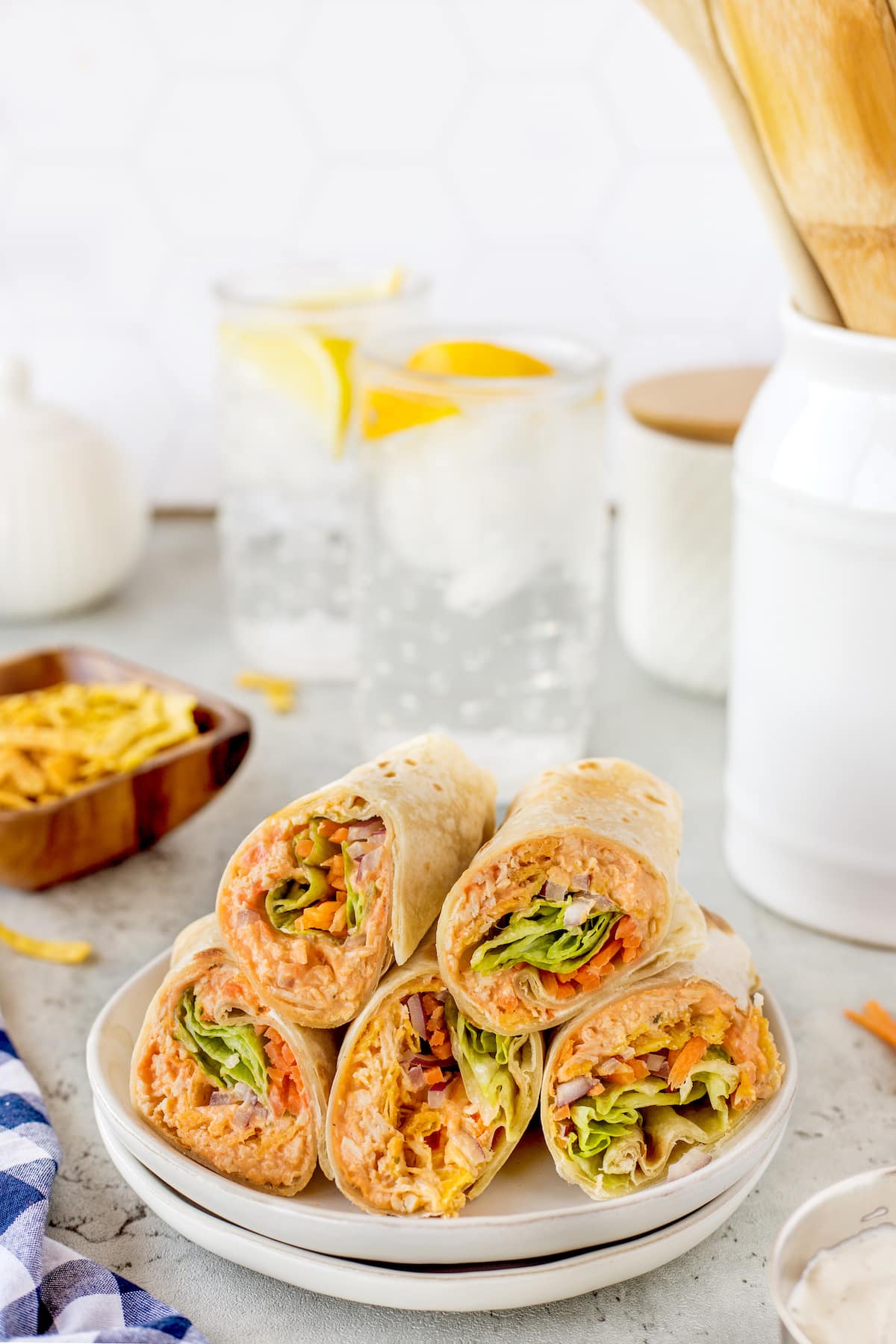 A plate of neatly stacked wrap halves filled with vegetables and meat. In the background, a glass of water with lemon slices, a wooden utensil holder, and decorative kitchen items are visible on a light-colored countertop.