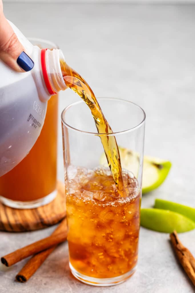 A hand pours apple cider from a jug into a glass filled with ice. Green apple slices and cinnamon sticks are on the table, set against a neutral background.