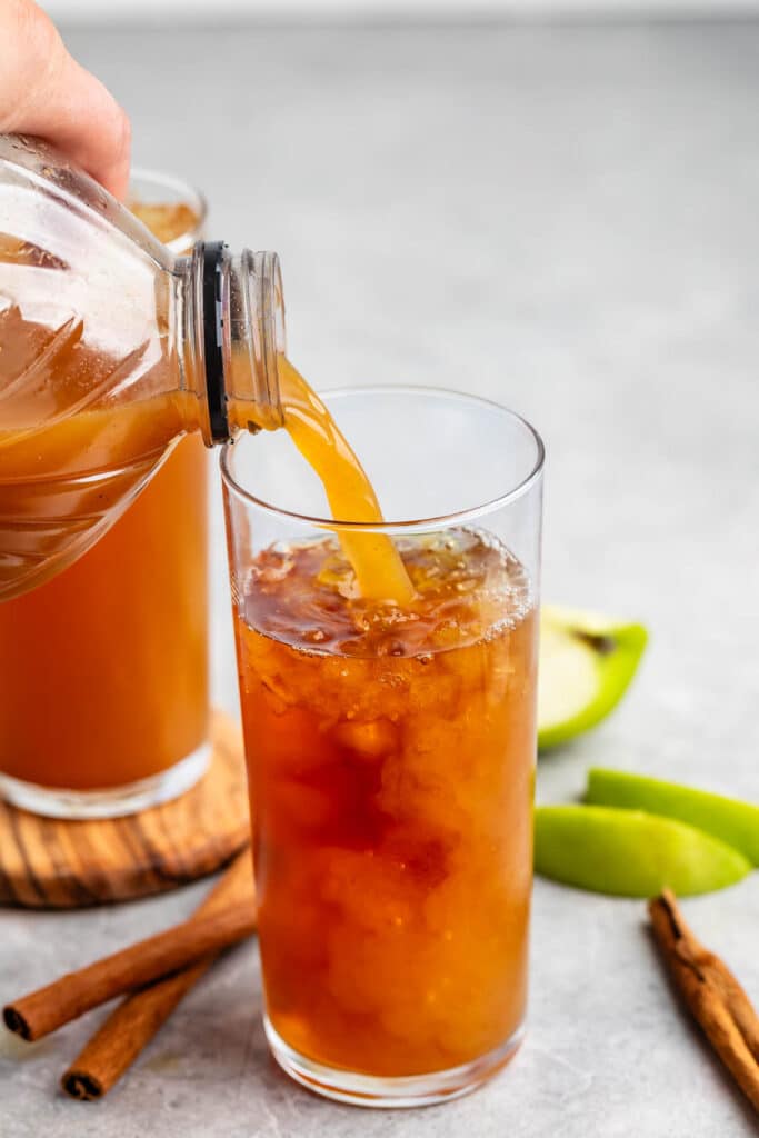 A bottle is pouring apple cider into a tall glass filled with ice. The drink is a warm amber color. In the background, cinnamon sticks and sliced green apples are on a gray surface. Another glass of cider is partially visible.