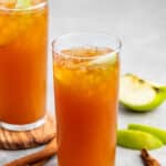 Two tall glasses filled with a brown, ice-cold beverage are garnished with apple slices. Cinnamon sticks are placed beside the glasses on a gray surface, and fresh apple slices are visible in the background.