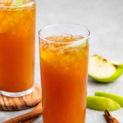 Two tall glasses filled with a brown, ice-cold beverage are garnished with apple slices. Cinnamon sticks are placed beside the glasses on a gray surface, and fresh apple slices are visible in the background.