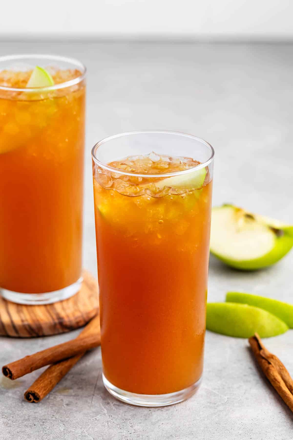 Two tall glasses filled with a brown, ice-cold beverage are garnished with apple slices. Cinnamon sticks are placed beside the glasses on a gray surface, and fresh apple slices are visible in the background.