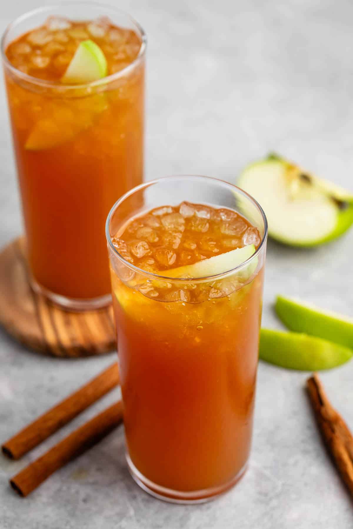 Two tall glasses of iced apple cider topped with apple slices, set on a light surface. Nearby are cinnamon sticks and apple slices, adding a touch of garnish and flavor.