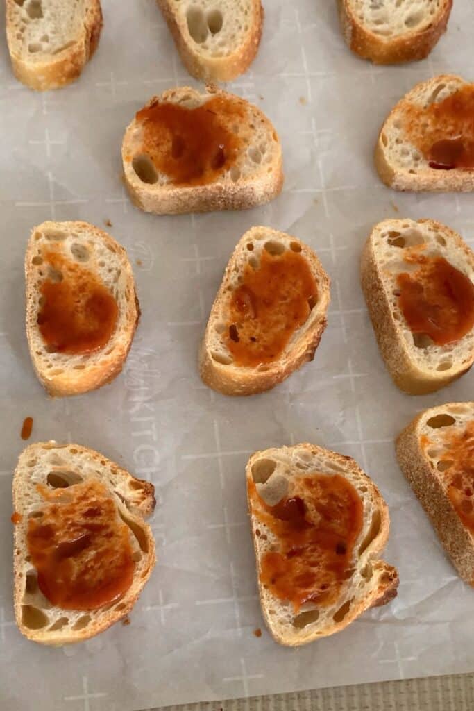 Slices of baguette topped with tomato sauce are arranged on parchment paper. The bread is lightly toasted and evenly spaced across the sheet.