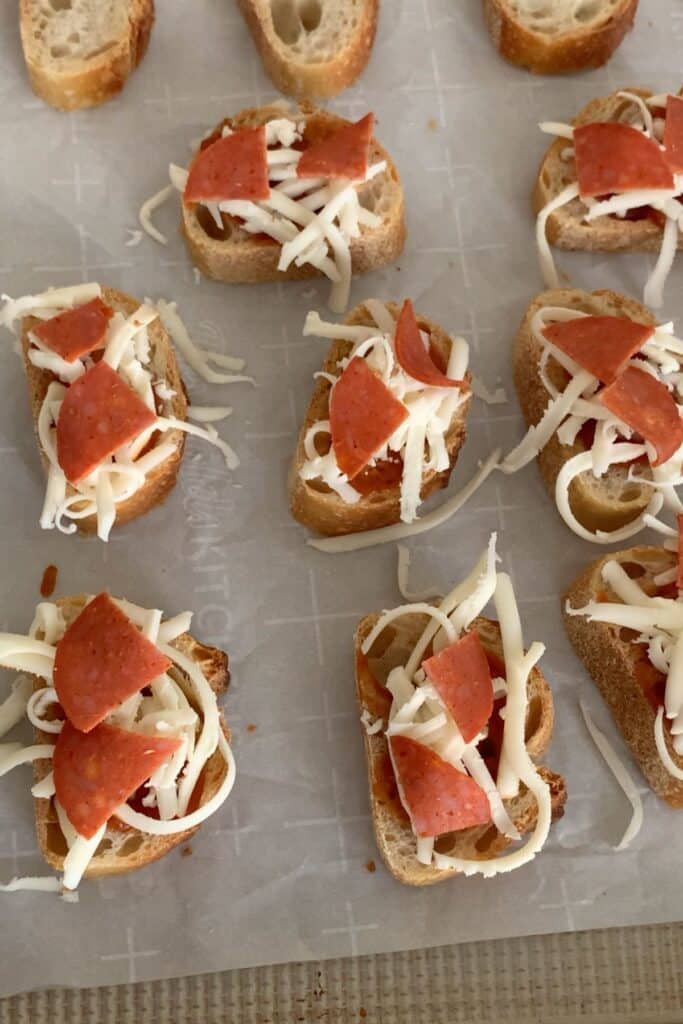 Slices of bread topped with shredded cheese and pepperoni are arranged on a parchment-lined baking sheet, ready to be baked.