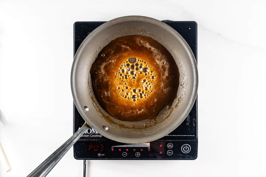 A steel pan on an induction cooktop filled with bubbling caramel sauce. The sauce is a rich brown color, and the cooktop display reads PO3.