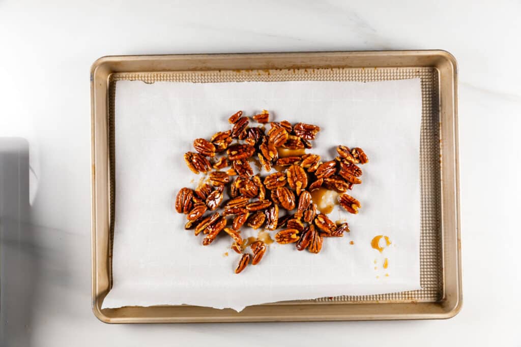 A baking tray lined with parchment paper holds a scattered pile of glazed pecans on a light-colored surface. The pecans appear roasted and coated with a shiny, sticky glaze.