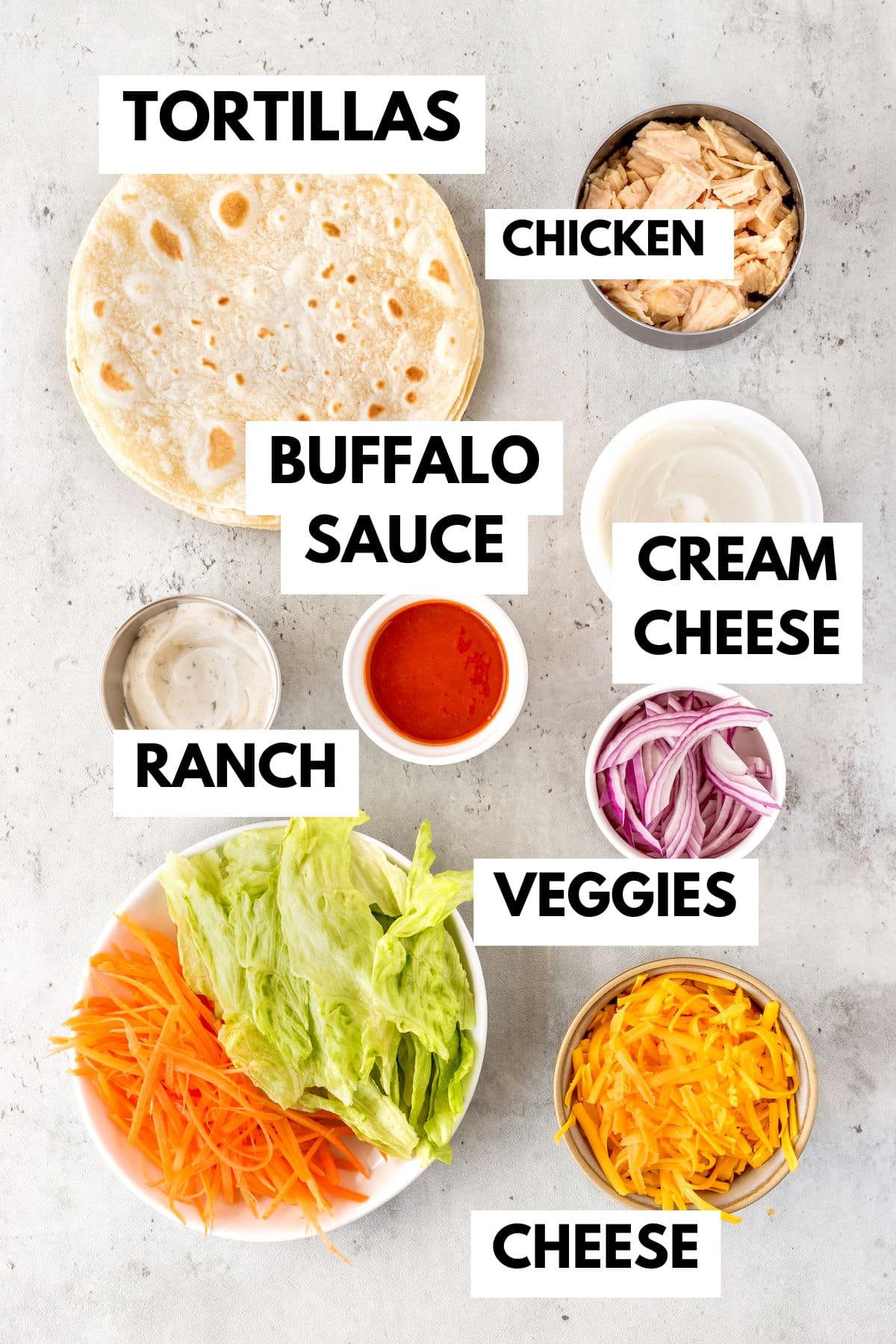 Ingredients neatly arranged on a light surface: tortillas, shredded chicken, buffalo sauce, cream cheese, ranch dressing, shredded cheese, julienned red onions, shredded lettuce, and carrots labeled in bowls and containers.