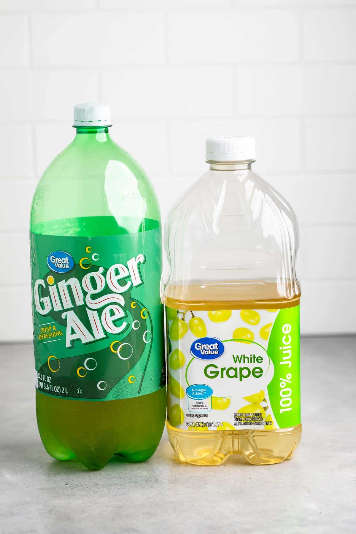 A green bottle of ginger ale and a clear bottle of white grape juice stand side by side on a gray countertop, with a white tiled wall in the background.