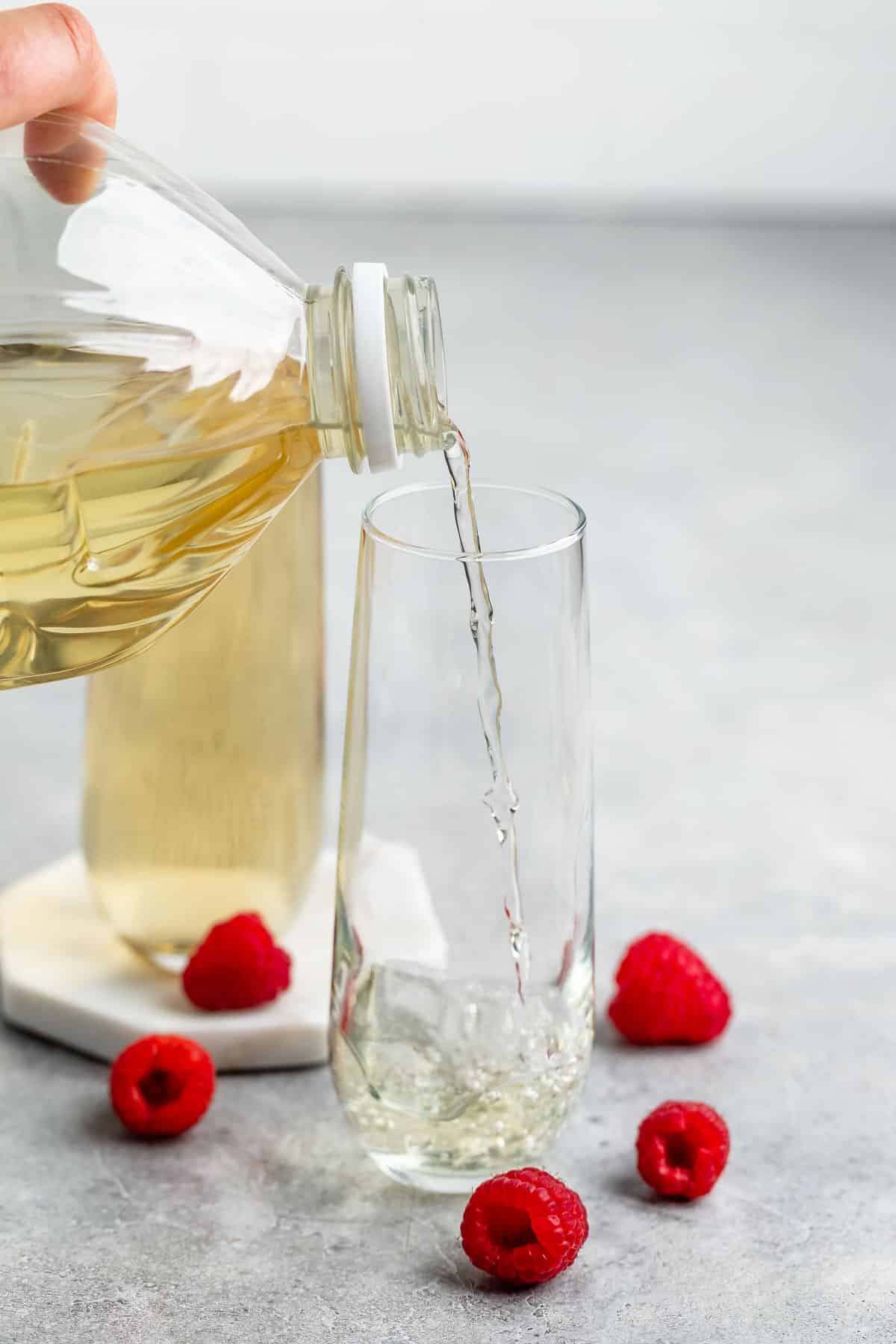 A hand pours a light-colored liquid into a tall, empty glass on a gray surface. Fresh raspberries are scattered around the glass, adding a pop of red color to the scene.