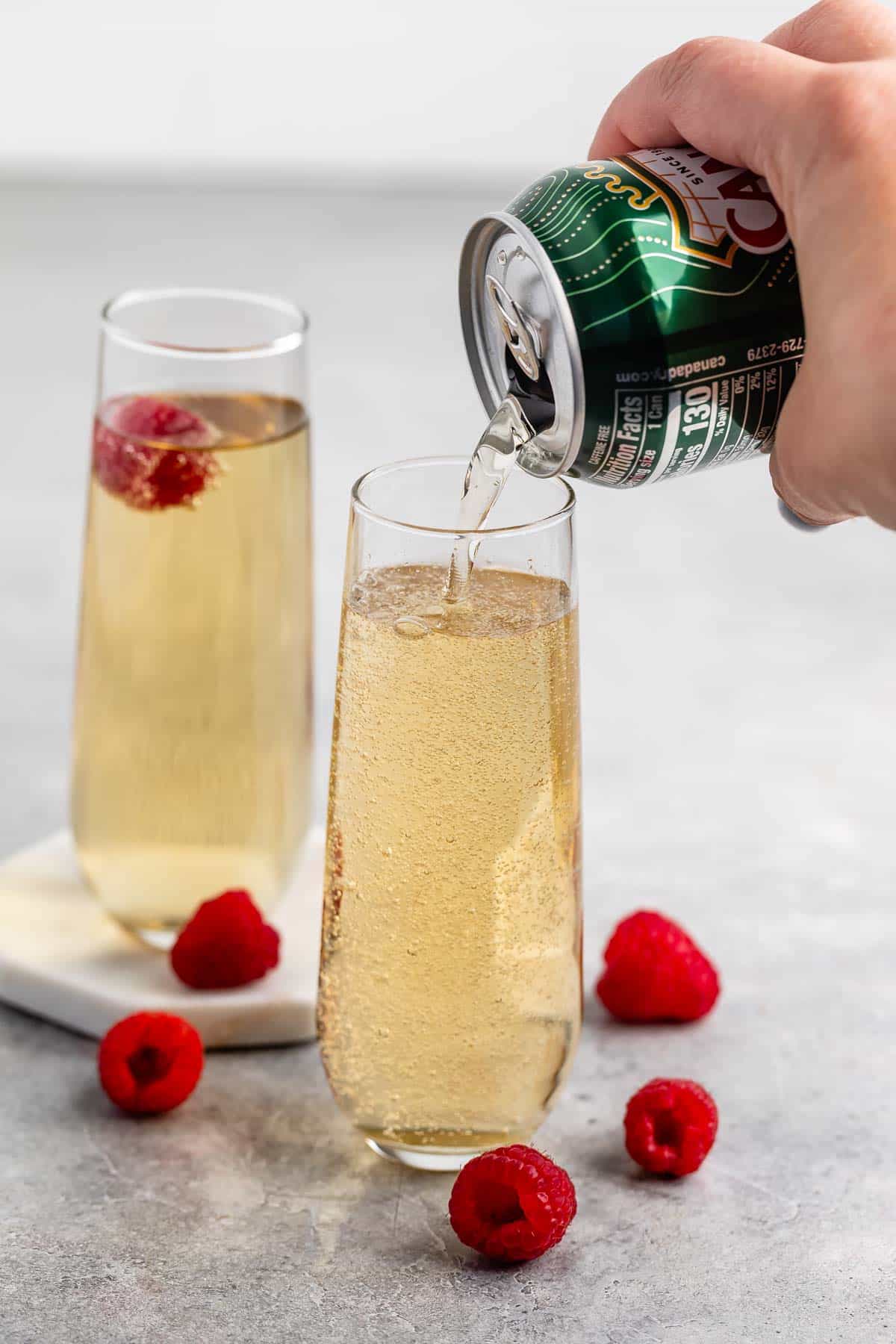 A can of ginger ale is being poured into a champagne flute. Another filled flute and several raspberries are in the background on a light gray surface.