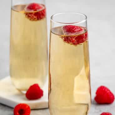 Two tall glasses of champagne are garnished with fresh raspberries. Additional raspberries are scattered on a light gray surface around the glasses, creating a refreshing and elegant scene.