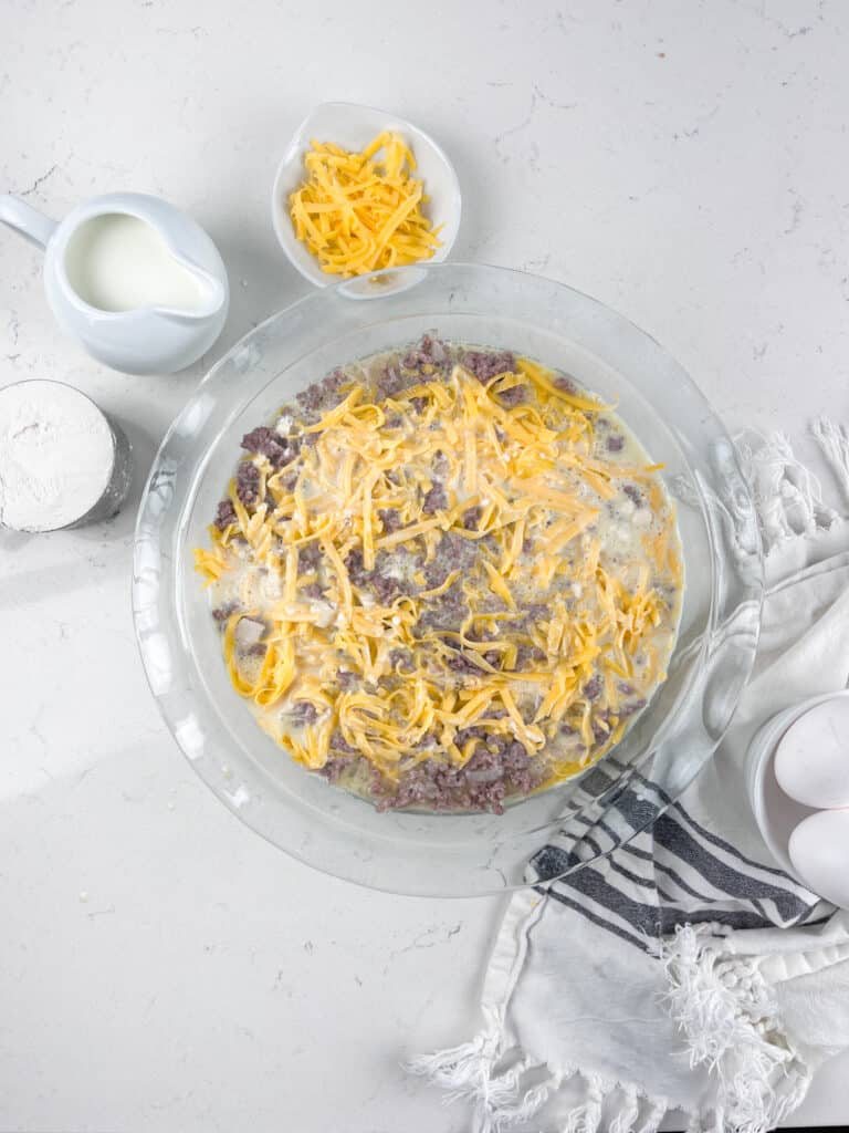 A glass baking dish on a white countertop contains a mixture of ground meat and shredded cheese. Nearby are a small pitcher of milk, a bowl of flour, more shredded cheese in a dish, a striped cloth, and two eggs.