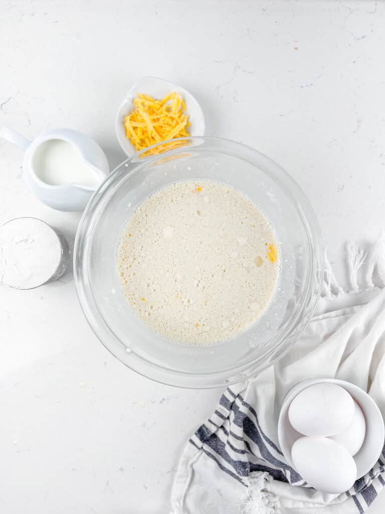 A glass bowl with a bubbly liquid mixture, surrounded by a jug of milk, a small bowl of shredded cheese, eggs in a bowl, and a container of flour on a white surface. A striped cloth napkin is nearby.