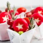 Brightly colored cheese rounds with star-shaped cutouts are arranged in white paper cups. Each cup contains decorative rosemary sprigs and grape skewers, creating a festive and cheerful presentation.