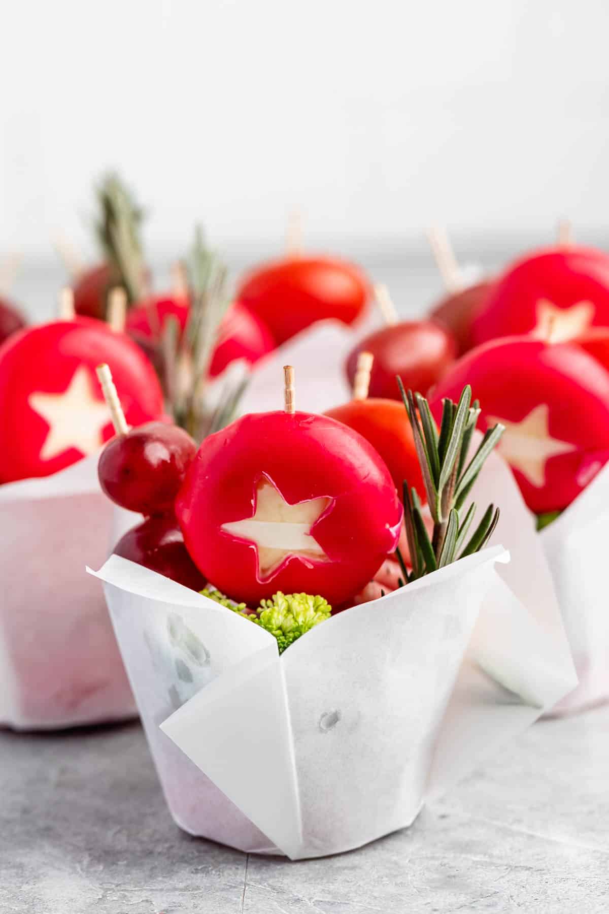 Brightly colored cheese rounds with star-shaped cutouts are arranged in white paper cups. Each cup contains decorative rosemary sprigs and grape skewers, creating a festive and cheerful presentation.