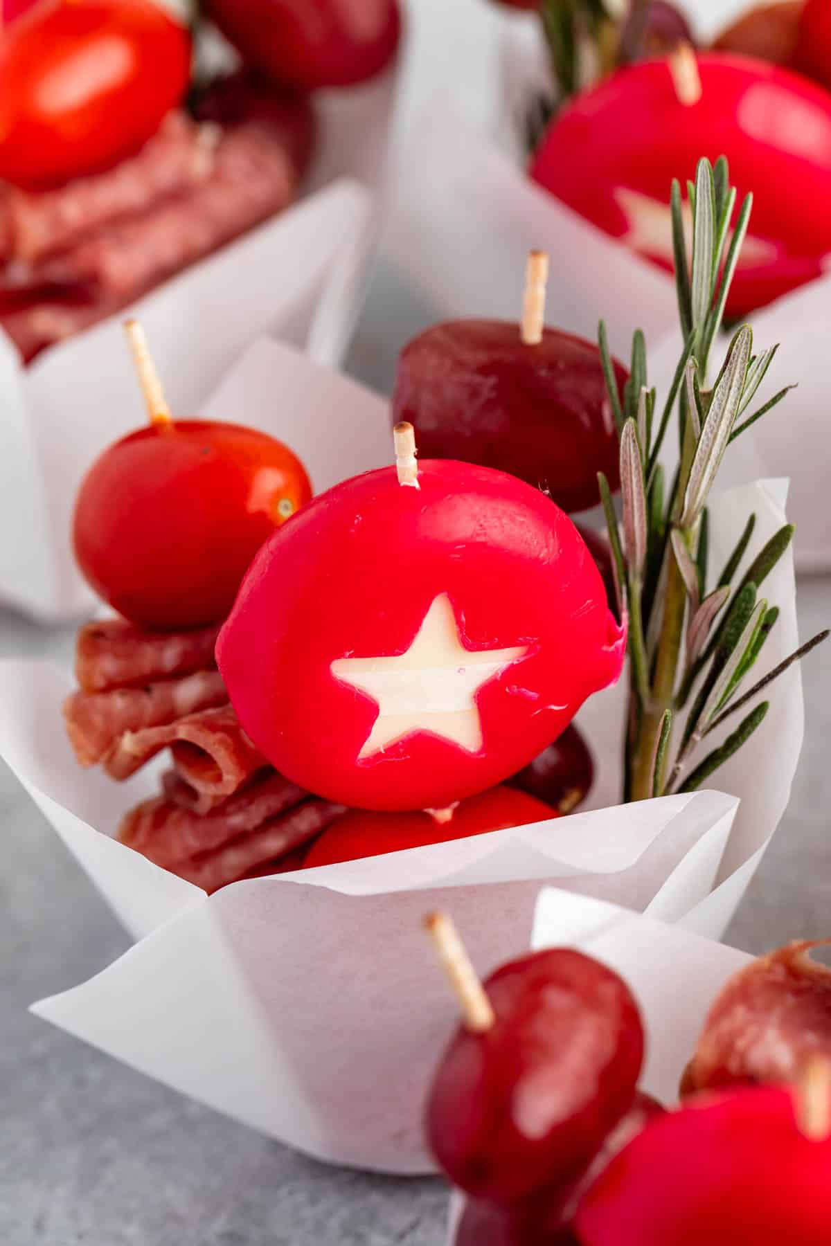 A close-up of a festive appetizer featuring a star-shaped cheese encased in red wax, with toothpicks holding cherry tomatoes, salami slices, and sprigs of rosemary, all arranged in a white paper cup.