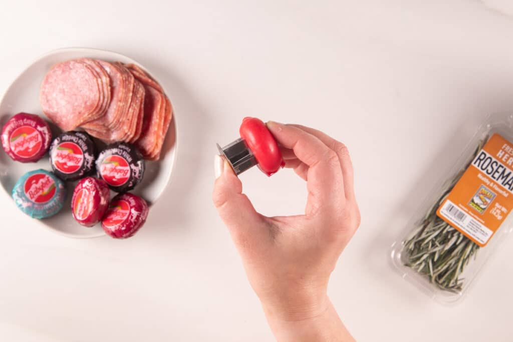 A hand holds a small red cheese wrapped in wax near a plate of various meats and cheeses. A package of fresh rosemary is on the right, laid out on a white countertop.