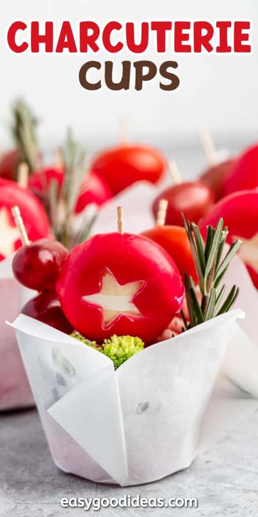 Close-up of a charcuterie cup featuring sliced star-shaped radishes, cherry tomatoes, broccoli, and rosemary sprigs. The cup is wrapped in white parchment paper, with the words Charcuterie Cups written above.