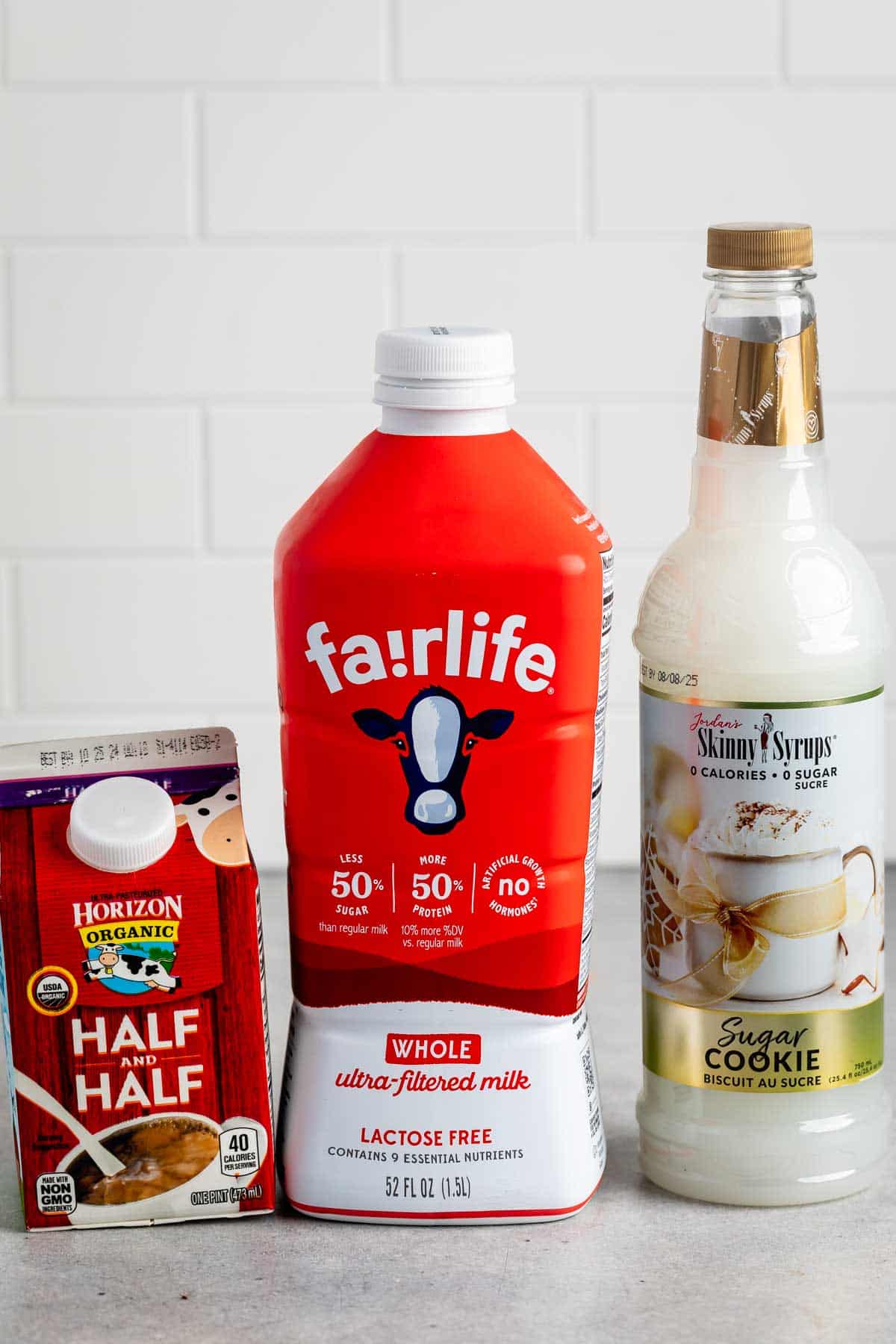 Three containers of coffee creamers are displayed: a carton of Horizon Organic Half and Half, a red bottle of Fairlife Whole Ultra-Filtered Milk, and a bottle of Skinny Syrups Sugar Cookie flavored syrup, set against a white tiled backdrop.