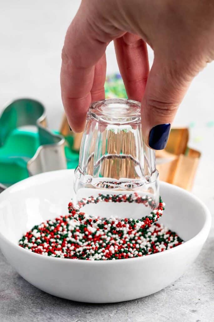 A hand holding an upside-down glass is dipping the rim into a white bowl filled with red, black, and white sprinkles. Assorted cookie cutters are in the background on a gray surface.