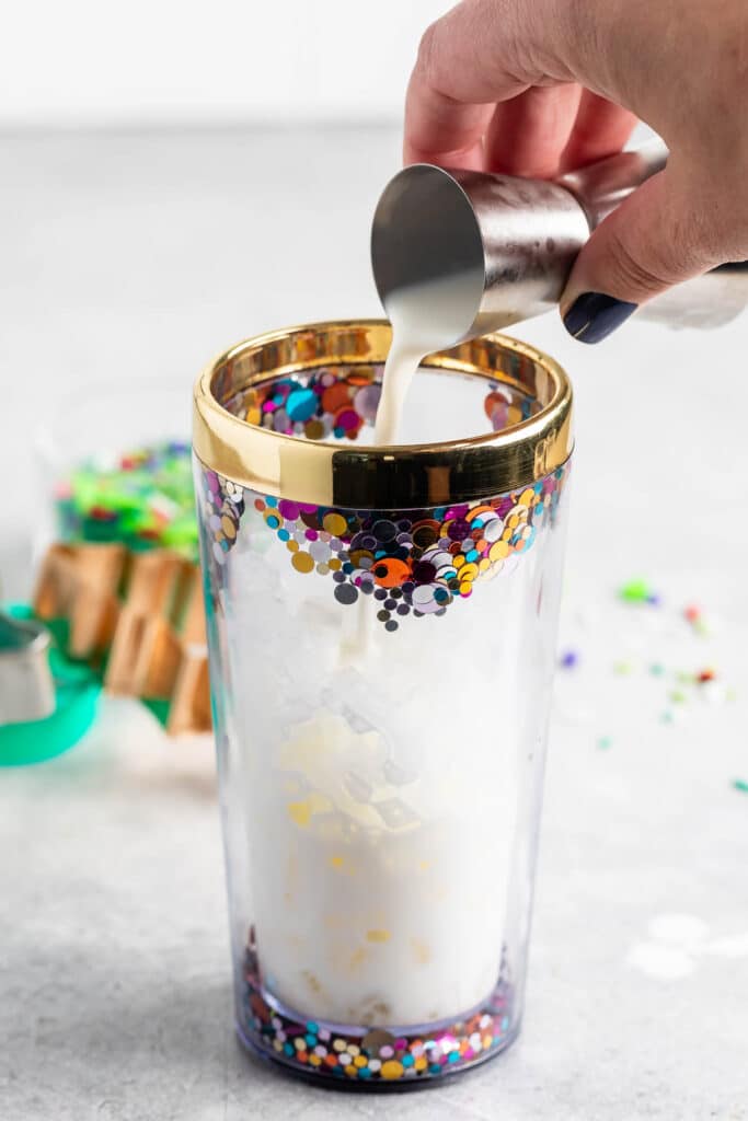 A hand holding a metal jigger pours a creamy liquid into a clear glass with gold trim and colorful confetti-like decorations. Background shows a blurred stack of similar glasses.